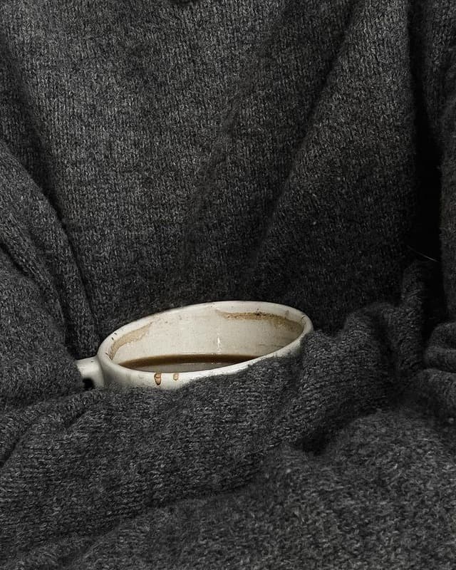 Hände in einem grauen Wollpullover, die eine Tasse Kaffee halten